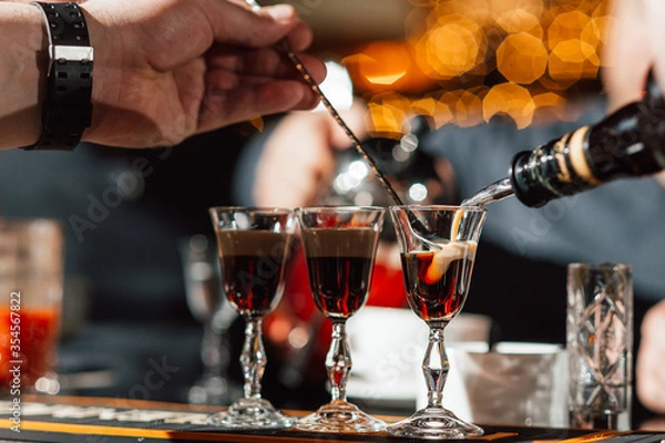 Fototapeta The bartender makes cocktails with liquor, using a bar spoon. layered cocktails. Bar counter. Atmospheric photo with warm tinting