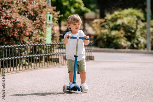 Obraz Boy learn to ride scooter in a park on sunny summer day. Preschooler boy riding a roller. Kids play outdoors with scooters.