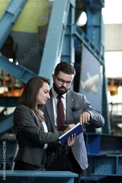 Obraz Portrait of a solid businessman with his secretary holding blue notebook,  talking about factory financial report in a airplane manufactory. Factory managers controlling work process in a factory