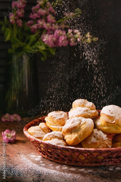 Fototapeta Homemade profiteroles with vanilla cream in a basket, rustic wooden table, sunny light in the kitchen, dark background with flowers, Selective focus, copy space. French choux pastry balls with cheese.