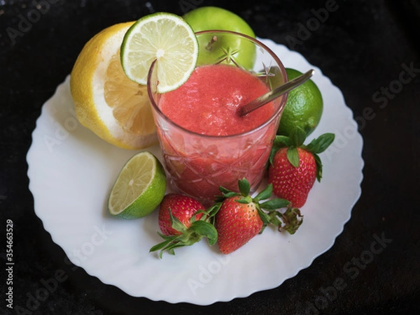 Fototapeta A glass of red cocktail with fruit on the white plate and dark background. Apple, lime, lemon, strawberry. Spoon inside the glass. 