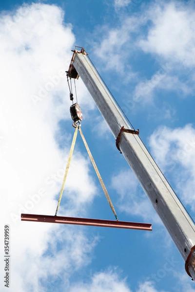 Fototapeta Construction crane with a hook and a metal profile hanging on ropes, low angle view with cloudy sunny blue sky