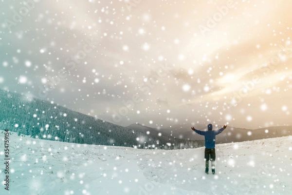 Fototapeta Back view of tourist hiker in warm clothing with backpack standing with raised arms on clearing covered with snow on spruce forest mountain and cloudy sky copy space background.