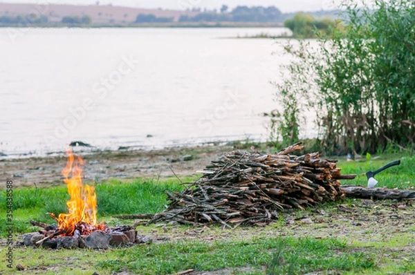 Obraz Small campfire with flames beside a lake with wood pile on the side.