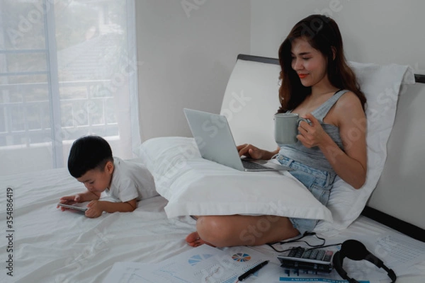 Obraz The mother works with her son in the bedroom at home. work at home, concept with a women sitting on the bed with laptop and holding a coffee mug.