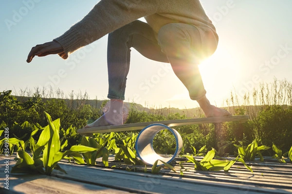 Fototapeta side view of healthy beautiful active woman keeping balance on the wooden balance board against the sky background at sunset summer day. Exercise and outdoor concept.