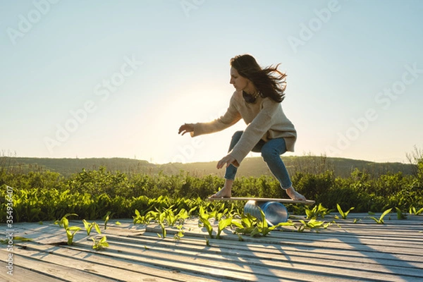 Fototapeta side view of healthy beautiful active woman keeping balance on the wooden balance board against the sky background at sunset summer day. Training and outdoor concept.