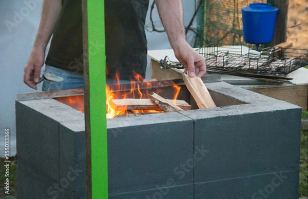 Fototapeta A man throws dry branches into the grill fire. Preparing fire for barbeque meat