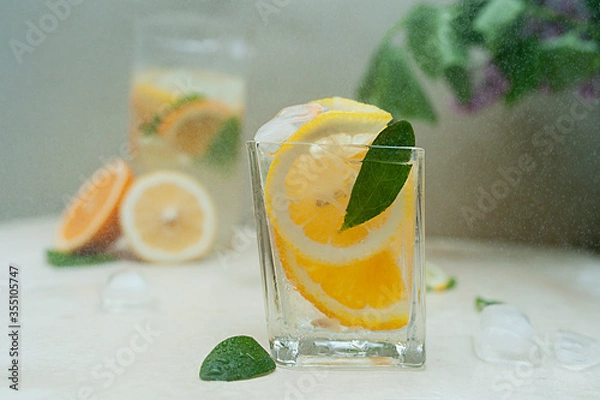 Fototapeta drops of water fly on a small wet glass with drops from condensation full of citrus fruits lemons and oranges with mint leaf and ice. homemade lemonade, online recipes