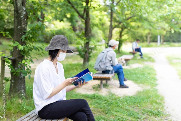 Fototapeta 公園のベンチで帽子をかぶりマスクをして読書する1人の女性