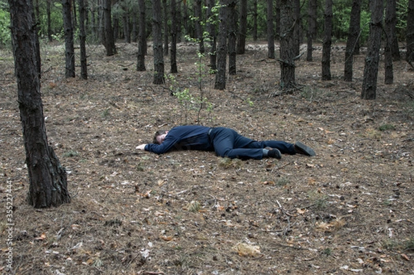 Obraz Murder in the woods. The body of a man in a blue shirt and trousers lies on the ground among the trees in the forest. Victim of an attack.