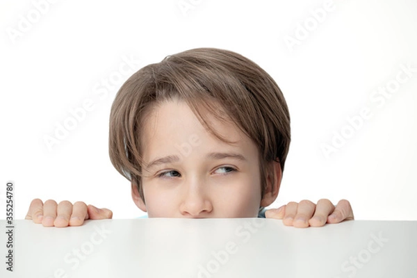 Fototapeta   boy peeping from behind a white table, the  portrait  of boy looks out, a sly look,  free copy space,