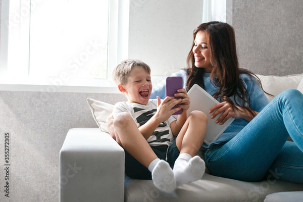 Fototapeta Happy young mother sit on couch using tablet relax with preschooler son holding smartphone have fun together