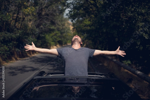 Fototapeta Young happy man drives a car and holds his hand out from the window. Driver enjoys driving and shows Ok sign with his hand out of window. Road trip, travel and freedom concept.