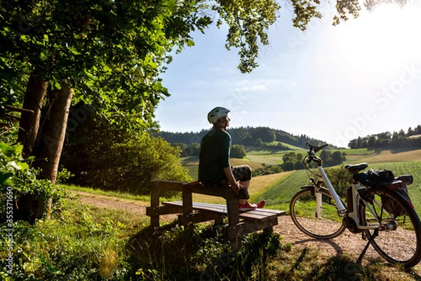 Fototapeta Frau genießt nach anstrengender Fahrradtour mit Ihrem Elektro Fahrrad die Abendsonne auf einer Holzbank in Idyllischer Hügellandschaft