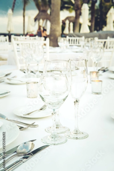 Fototapeta Festive serving with glasses for an outdoor celebration with garland lamps and palms at the background. Wedding, anniversary, birthday dinner and party. Toned image with place for text