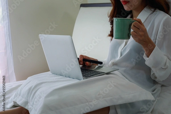 Obraz Relaxing work at home, concept with a women sitting on the bed with laptop and holding a coffee mug.