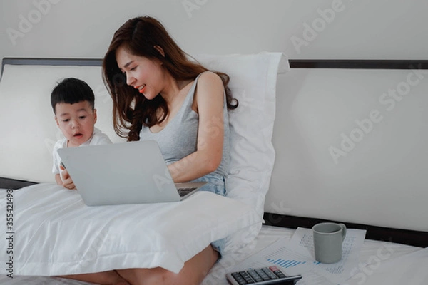 Obraz The mother works with her son in the bedroom at home. work at home, concept with a women sitting on the bed with laptop and holding a coffee mug.