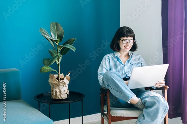 Obraz Beautiful brunette with a short haircut works in the home office on a blue background. Typing on a laptop, calling on the phone, transferring documents, making notes, drinking coffee from a mug.