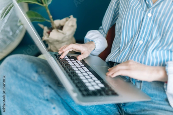 Obraz Beautiful brunette with a short haircut works in the home office on a blue background. Typing on a laptop, calling on the phone, transferring documents, making notes, drinking coffee from a mug.