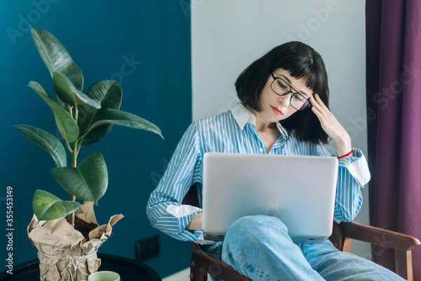 Obraz Beautiful brunette with a short haircut works in the home office on a blue background. Typing on a laptop, calling on the phone, transferring documents, making notes, drinking coffee from a mug.