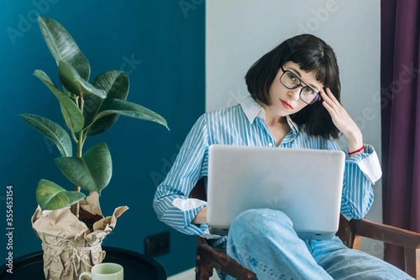 Obraz Beautiful brunette with a short haircut works in the home office on a blue background. Typing on a laptop, calling on the phone, transferring documents, making notes, drinking coffee from a mug.