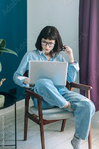 Obraz Beautiful brunette with a short haircut works in the home office on a blue background. Typing on a laptop, calling on the phone, transferring documents, making notes, drinking coffee from a mug.