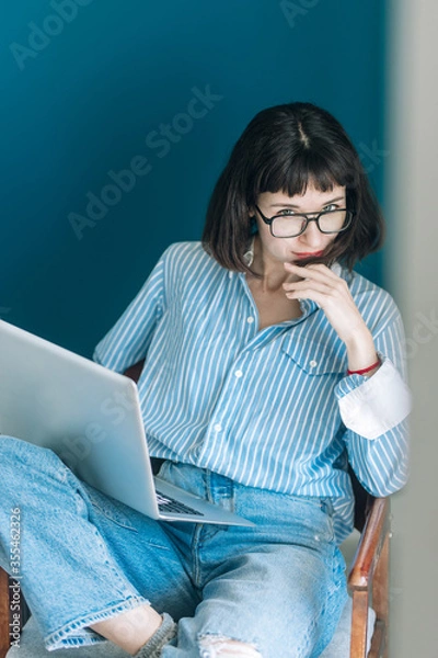 Obraz Beautiful brunette with a short haircut works in the home office on a blue background. Typing on a laptop, calling on the phone, transferring documents, making notes, drinking coffee from a mug.