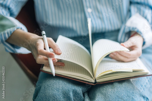 Obraz Beautiful brunette with a short haircut works in the home office on a blue background. Typing on a laptop, calling on the phone, transferring documents, making notes, drinking coffee from a mug.
