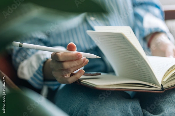 Obraz Beautiful brunette with a short haircut works in the home office on a blue background. Typing on a laptop, calling on the phone, transferring documents, making notes, drinking coffee from a mug.