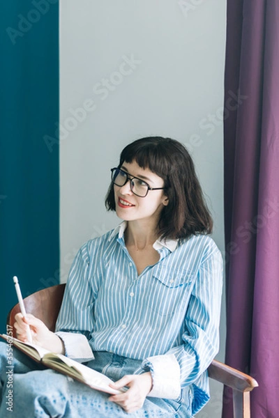 Obraz Beautiful brunette with a short haircut works in the home office on a blue background. Typing on a laptop, calling on the phone, transferring documents, making notes, drinking coffee from a mug.