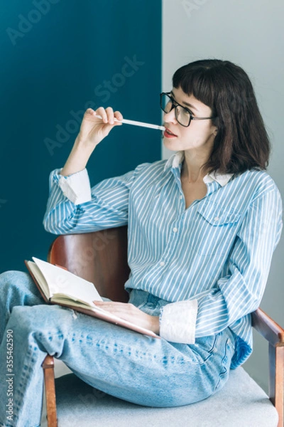 Obraz Beautiful brunette with a short haircut works in the home office on a blue background. Typing on a laptop, calling on the phone, transferring documents, making notes, drinking coffee from a mug.