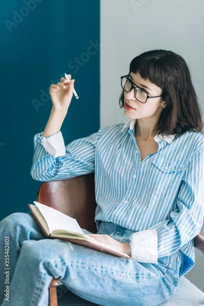 Obraz Beautiful brunette with a short haircut works in the home office on a blue background. Typing on a laptop, calling on the phone, transferring documents, making notes, drinking coffee from a mug.