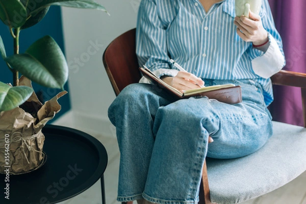 Obraz Beautiful brunette with a short haircut works in the home office on a blue background. Typing on a laptop, calling on the phone, transferring documents, making notes, drinking coffee from a mug.