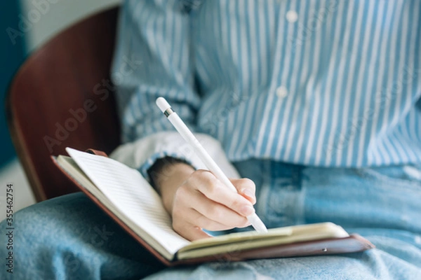 Obraz Beautiful brunette with a short haircut works in the home office on a blue background. Typing on a laptop, calling on the phone, transferring documents, making notes, drinking coffee from a mug.