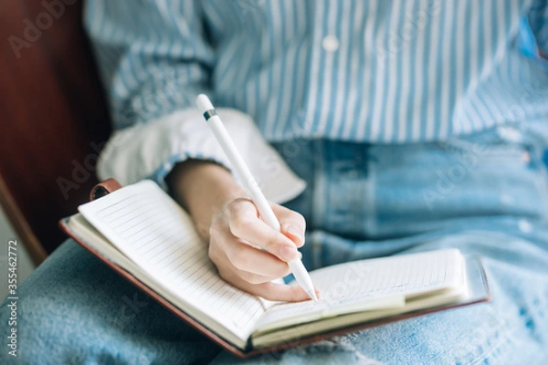 Obraz Beautiful brunette with a short haircut works in the home office on a blue background. Typing on a laptop, calling on the phone, transferring documents, making notes, drinking coffee from a mug.