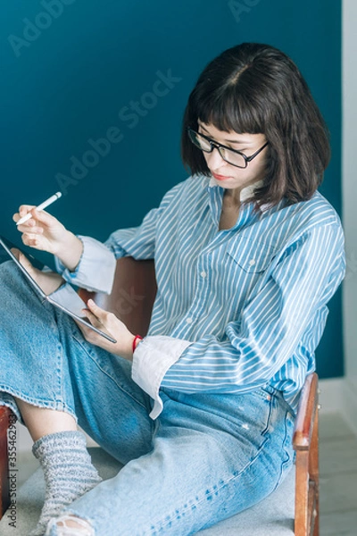 Obraz Beautiful brunette with a short haircut works in the home office on a blue background. Typing on a laptop, calling on the phone, transferring documents, making notes, drinking coffee from a mug.