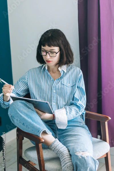 Obraz Beautiful brunette with a short haircut works in the home office on a blue background. Typing on a laptop, calling on the phone, transferring documents, making notes, drinking coffee from a mug.