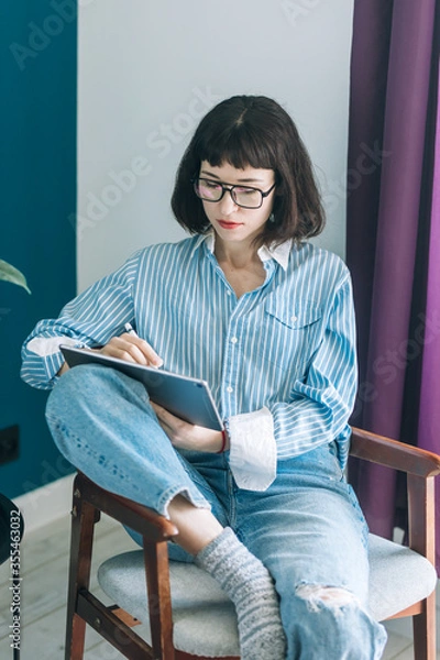 Obraz Beautiful brunette with a short haircut works in the home office on a blue background. Typing on a laptop, calling on the phone, transferring documents, making notes, drinking coffee from a mug.