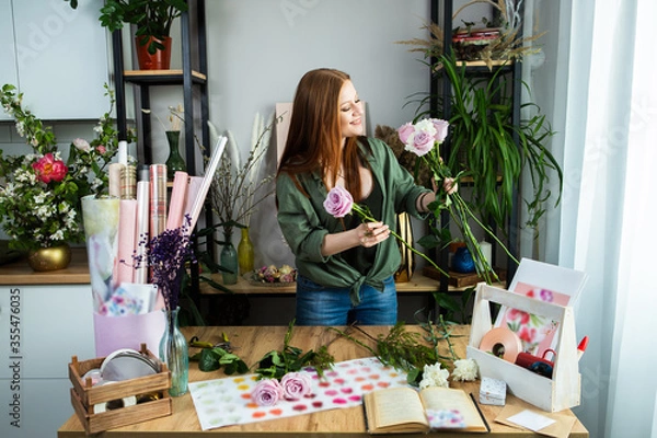 Fototapeta A girl florist with red hair collects a bouquet of roses in a flower shop. Remote workshop for the Assembly of bouquets.