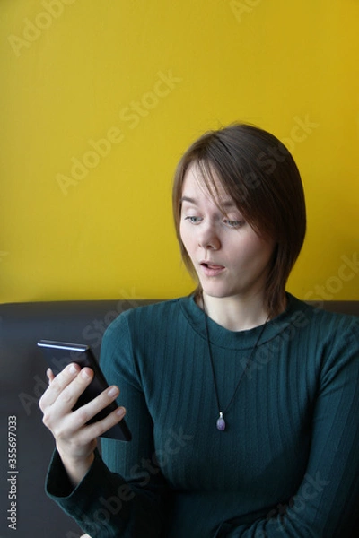 Fototapeta A young beautiful girl of European appearance in a blue jacket sits in a cafe with a mobile phone on the couch against the background of a yellow wall. Surprised expression. Place for text, copy space