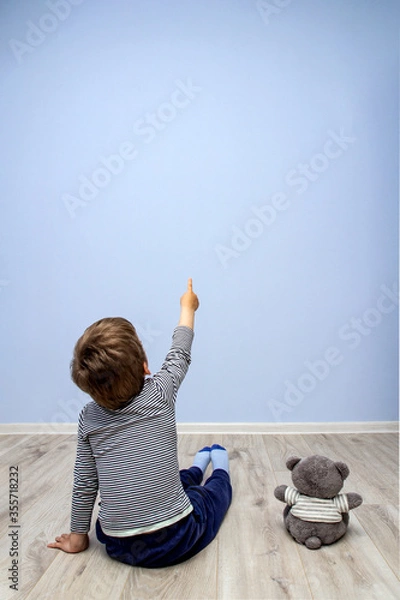 Obraz a child sitting on the floor with a bear cub points at a blue textured wall. copy space