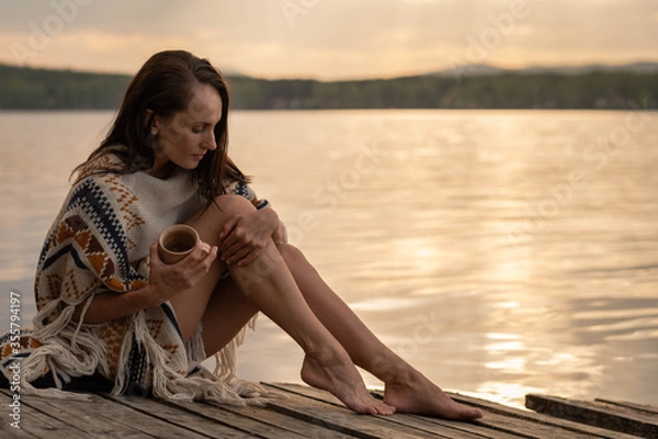 Obraz A beautiful dark-haired woman sits on an old wooden pier in the early hours of dawn holding a ceramic mug of fragrant tea, stroking her leg