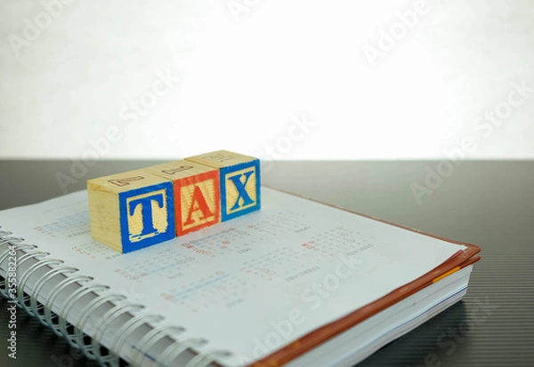 Fototapeta Conceptual image of annual tax return.  Wooden block letter forming TAX on note book over dark reflective surface. Focus at block letter A.