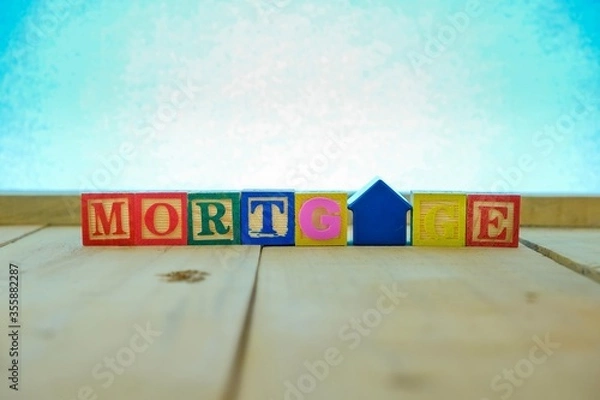 Fototapeta Conceptual of mortgage and home loan. Wooden block letters and  house-shape key chain hand held spelling the words MORTGAGE on wooden table. Focus at blue letter T. Bluish background.