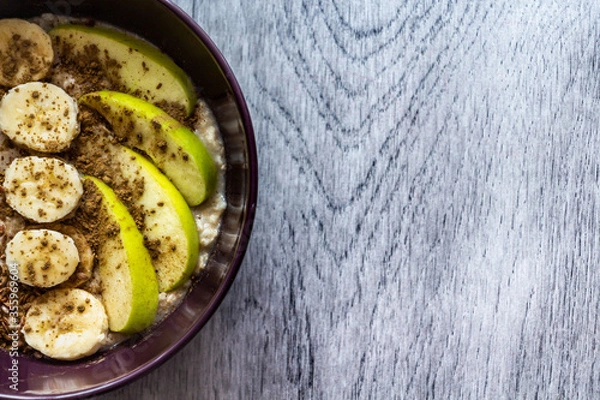 Obraz Plate of oatmeal with apple, banana, cinnamon and flax seeds stands on a gray wooden background. Top view. Copy space for text. Healthy nutrition concept