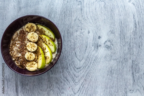 Obraz Plate of oatmeal with apple, banana, cinnamon and flax seeds stands on a gray wooden background. Top view. Copy space for text. Healthy breakfast concept