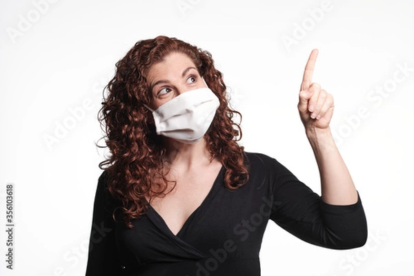 Fototapeta Redhead woman wearing a protective mask for coronavirus and pointing up with her finger