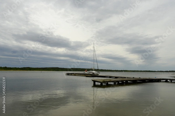 Fototapeta A view of an old wooden jetty, marina, or platform with a single boat without a sail floating next to it with a cloudy spring sky before the storm reflecting in the surface of the lake or river