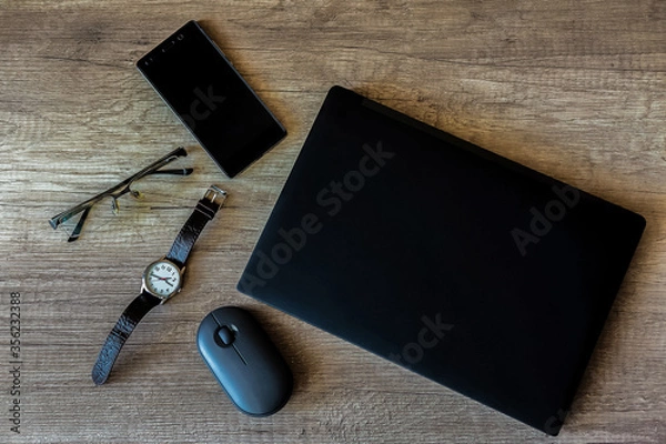 Obraz On a textured brown wooden table is a home work space with a black laptop, mobile phone, wristwatch, glasses, and computer mouse. Close-up view from above.